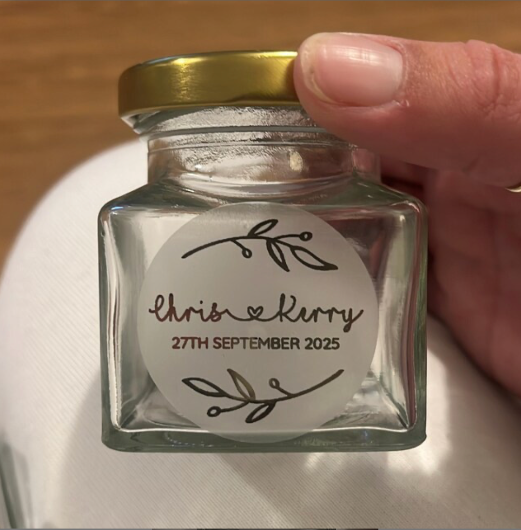 Customer's hand holding up a small jam jar wedding favour with personalised rustic leaves wedding sticker