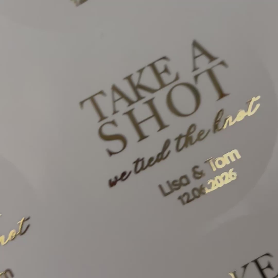 Load video: Personalised 'take shot, we've tied the knot' Wedding favour Stickers moving to show the high shine gold foil finish